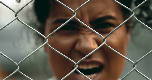 Eine aufgebrachte junge schwarze Frau schreit hinter Metallzäunen und blickt in die Kamera, die vor Wut grimassiert und vor Verzweiflung schreit. Zwanziger empfindet Empörung - Foto, Bild