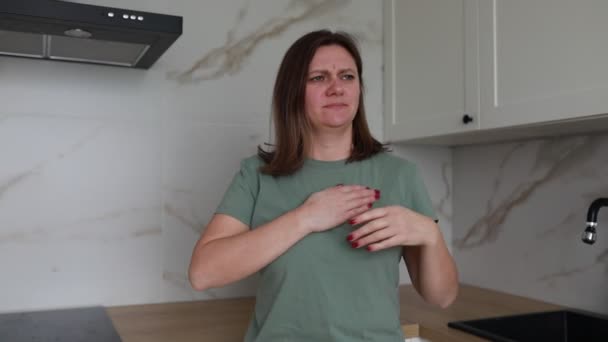 Доросла жінка стоїть на сучасній кухні з болючим виразом, стискаючи груди в дискомфорті, що може свідчити про проблеми зі здоров'ям, такі як печія або потенційний серцевий біль. Зрозуміло - Кадри, відео