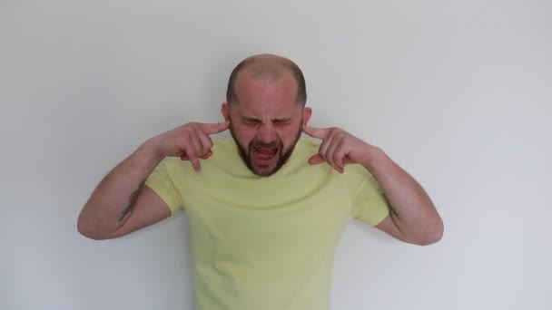 A bald man wearing a yellow t-shirt stands against a white background, dramatically covering his ears with his fingers and grimacing, suggesting he is trying to block out a loud sound or showing a - Footage, Video