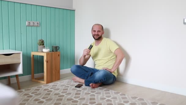 A cheerful, bald man sits cross-legged on a tiled living room floor against a teal backdrop, savoring a chocolate ice cream bar. With a warm smile and casual attire, he seems to be enjoying a - Footage, Video
