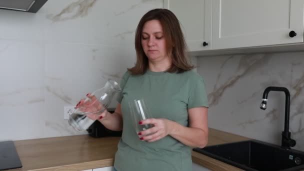 Egy nő megtölt egy poharat vízzel egy tiszta kancsóból, a konyhája világos, kortárs környezetében. A márvány backsplash és rozsdamentes acél mosogató hangsúlyozza a modern design elemek. - Felvétel, videó