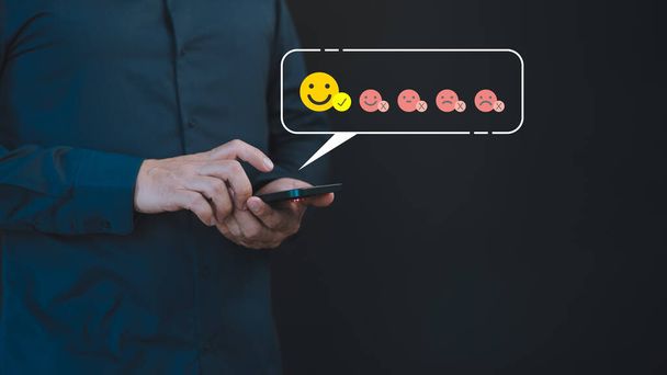 A male hand uses a mobile phone displaying a happy smile face icon to express satisfaction with the service, indicating a very impressed opinion rating. - Photo, Image