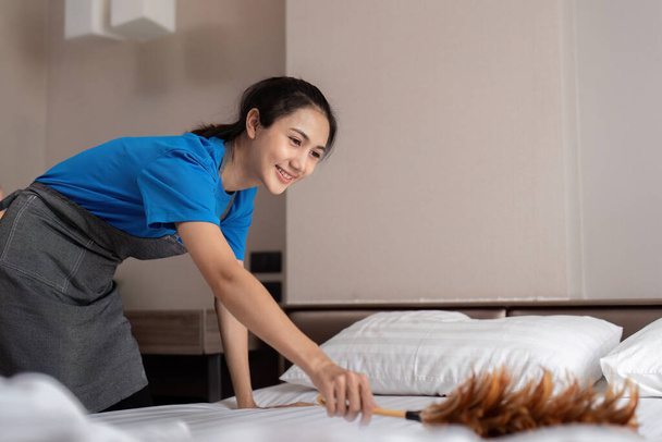 女性アジア人スタッフのクリーニングサービス,ツール,バケツ. 寝室をきれいにするプロダクトが付いている若い女性のクリーナー,ベッドから塵. - 写真・画像