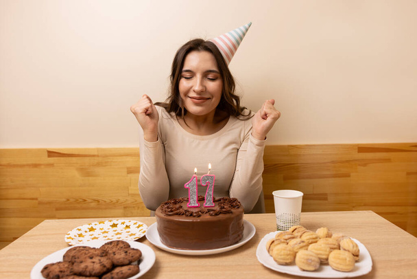 Ein Mädchen sitzt vor einem Tisch mit einer festlichen Torte, in der eine Kerze in Form der Zahl 17 entzündet wird. Das Konzept einer Geburtstagsfeier. - Foto, Bild