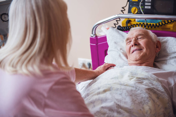 Веселый старик смотрит на свою жену и улыбается, лежа в постели в больничной палате
 - Фото, изображение