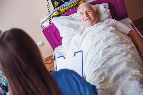 Bello vecchio sta parlando con un'infermiera e sorridente mentre si trova a letto nel reparto ospedaliero
 - Foto, immagini