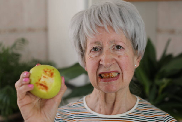 retrato de mulher de cabelos grisalhos maduros com dentes sangrando comer maçã verde em casa - Foto, Imagem