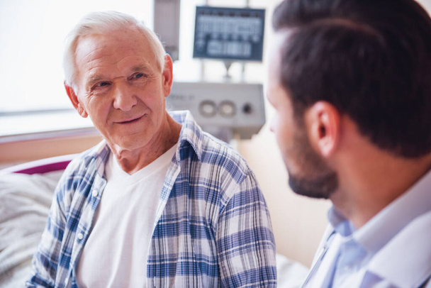 Knappe grijsaard is praten met een arts zittend in ziekenhuis ward - Foto, afbeelding