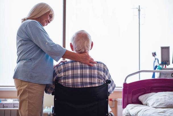 Красива стара леді погладжує чоловіка і посміхається, коли він сидить у інвалідному візку в лікарняному відділенні
 - Фото, зображення