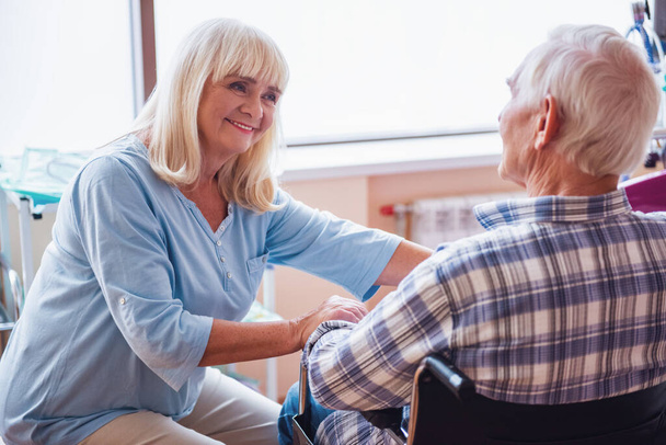 Bela senhora idosa está falando com seu marido e sorrindo enquanto ele está sentado na cadeira de rodas na enfermaria do hospital
 - Foto, Imagem