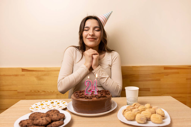 Dívka sedí před stolem se slavnostním dortem, ve kterém svíčka svítí v podobě čísla24. Koncept oslavy narozenin. - Fotografie, Obrázek