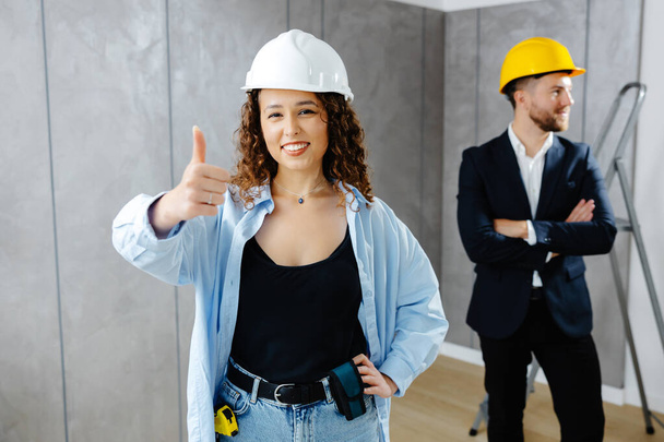 We've got the go ahead to renovate. Attractive young contractor and customer standing in room in helmet, looking at camera and showing gesture fingers up. - Photo, Image