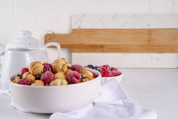 Крошечные круассаны на завтрак со свежими ягодами и молоком. Чаши с модной свежей выпечкой мини круассаны с малиной и черникой, десерт утренняя закуска - Фото, изображение