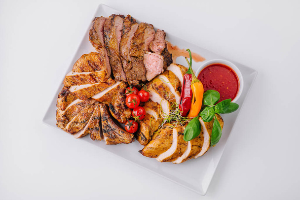 Bovenaanzicht van heerlijke gegrilde vleesschijfjes geserveerd met verse groenten en dipsaus op een wit bord - Foto, afbeelding