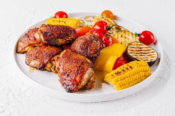 Παχιά μπούτια κοτόπουλου στη σχάρα μαζί με ψητό καλαμπόκι, πιπεριές, ντομάτες και μελιτζάνες σε λευκό πιάτο - Φωτογραφία, εικόνα