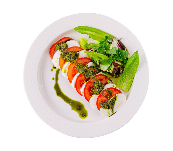 Bovenaanzicht van kapperssalade met rijpe tomaten, mozzarella, basilicum pesto, en groenten op witte plaat - Foto, afbeelding
