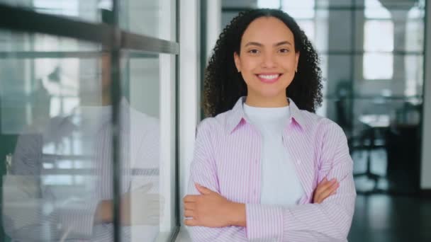 Female video portrait of a confident gorgeous successful hispanic or brazilian stylish curly haired young woman, stands in a modern office with arms crossed, looking at the camera, smiles friendly - Footage, Video