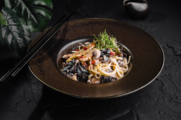 Gourmet roerbak noedels met zeevruchten, gegarneerd met microgreens, gepresenteerd op een stijlvolle donkere plaat - Foto, afbeelding