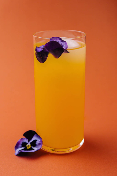 Levendige sinaasappel drank in een hoog glas, geaccentueerd met een delicate paarse eetbare bloem, op een warme achtergrond - Foto, afbeelding