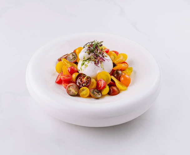 Свежая буррата с разноцветными помидорами черри и травами, представленная на белом блюде, мраморном фоне - Фото, изображение