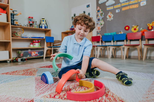 Маленький мальчик играет с радужной деревянной игрушкой, сидя на полу в детском саду. Селективный фокус. - Фото, изображение