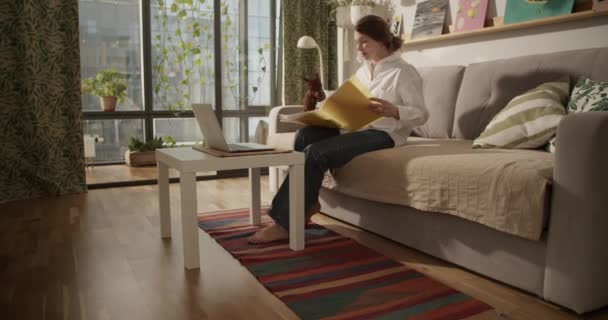 Real time side view of concentrated barefoot adult female κοιτάζοντας μακριά, ενώ εργάζονται από το σπίτι για laptop έγγραφα αναφοράς και παίζει με το κατοικίδιο ζώο σκυλί στον καναπέ  - Πλάνα, βίντεο