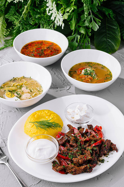 Verscheidenheid aan heerlijke soepen en een gegrilde vleesschotel, prachtig geserveerd op een getextureerde witte tafel, klaar om te eten - Foto, afbeelding