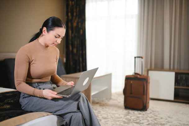 Young businesswoman working from hotel room on business trip, woman sitting on bed and using laptop. - Фото, изображение