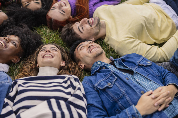 Ein enger Kreis junger Freunde liegt zurück auf dem Rasen und teilt die Freude in einem ehrlichen Moment des Lachens und der Entspannung.. - Foto, Bild