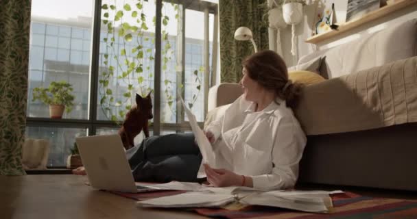 Widok w czasie rzeczywistym z boku skoncentrowany boso dorosła kobieta patrząc w dół na papierkową robotę leżąc na podłodze z laptopem karmienia i zabawy z psem podczas pracy z domu - Materiał filmowy, wideo