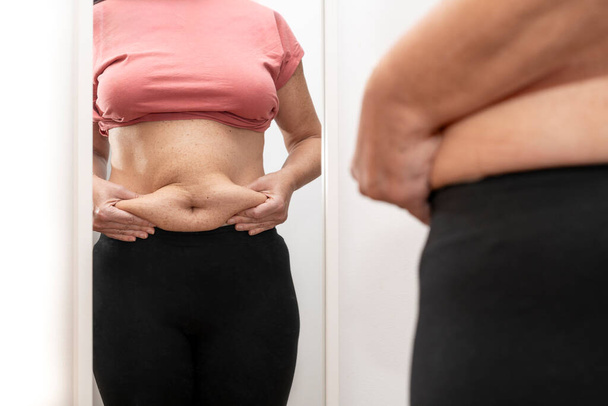 Primer plano de la mujer tomando exceso de grasa de su abdomen y mirándose en el espejo. Foto de alta calidad - Foto, imagen