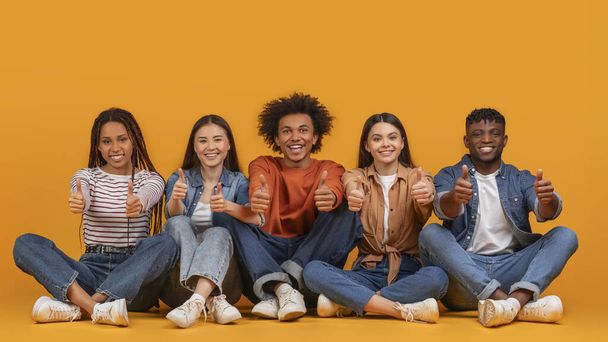 Egy sokszínű, öt barátból álló csapat feladja a hüvelykujját, miközben ül, jóváhagyást és barátságot mutat egy többnemzetiségű, többnemzetiségű, sárga alapon elszigetelt környezetben. - Fotó, kép
