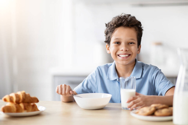 Een vrolijke Afro-Amerikaanse jongen wordt met zijn ontbijt, glimlachend, een uitnodigende sfeer van vreugde en gezinsleven in de keuken - Foto, afbeelding