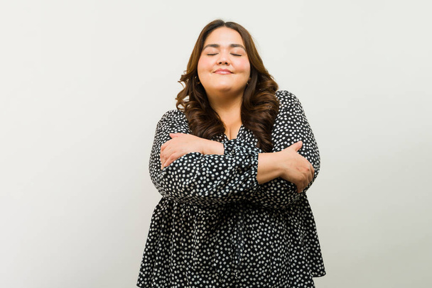 Прелестная plus-size женщина с обнимает себя в знак самоуважения в студии на белом фоне - Фото, изображение