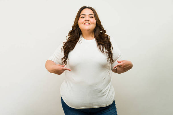 Mujer confiada de talla grande orgullosamente de pie en una camiseta blanca y pantalones vaqueros contra un telón de fondo neutral, sonriendo - Foto, imagen