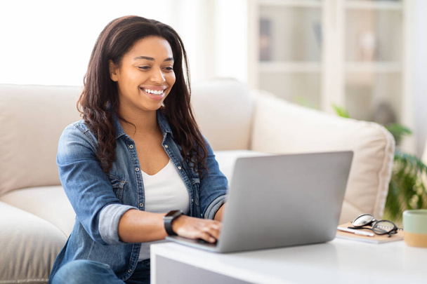 La empresaria afroamericana está sentada en un sofá, enfocada en usar una computadora portátil. Ella está escribiendo en el teclado y mirando la pantalla atentamente mientras trabaja o navega por Internet. - Foto, Imagen