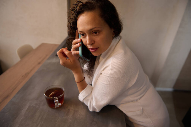 Joven mujer multiétnica mirando a un lado, hablando por teléfono móvil inteligente mientras toma el desayuno en casa, de pie en el mostrador de la cocina en casa, vestido con albornoz blanco. Retrato lateral de primer plano - Foto, Imagen