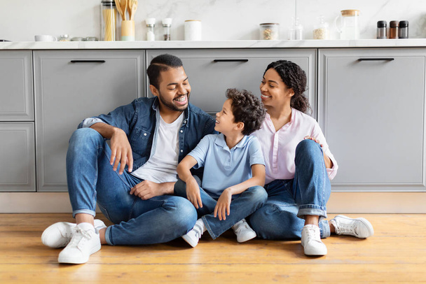 Neşeli bir Afro-Amerikan ailesi, modern bir mutfak zemininde oturmuş ailevi mutluluğu temsil ederken gülüşlerini paylaşıyor. - Fotoğraf, Görsel