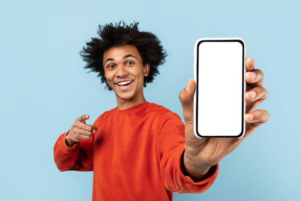 Energetischer afrikanisch-amerikanischer Zoomer zeigt auf einen leeren Smartphone-Bildschirm, ideal für App-Werbung, vor einem isolierten blauen Hintergrund - Foto, Bild