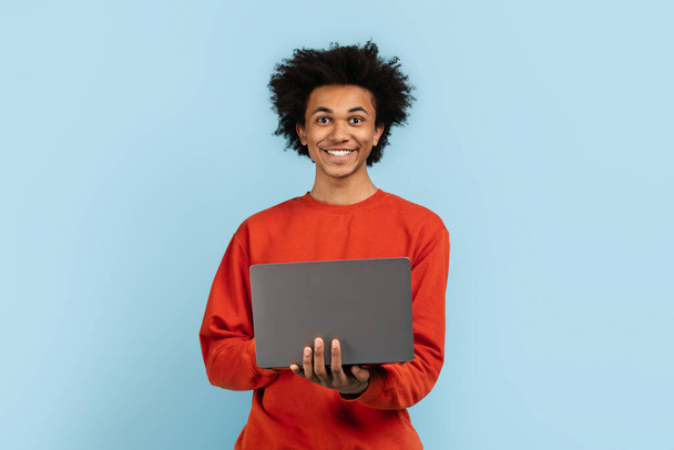 Elinde dizüstü bilgisayar tutan içerikli Afro-Amerikan bir adam, mavi arka planda izole edilmiş, teknolojiyle tatmin olmuş. - Fotoğraf, Görsel