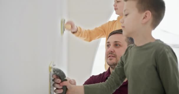 Father with his two sons engaging in a DIY home renovation project. They collaboratively paint the walls of their new home, sharing smiles and creating memories in a bright, modern space. - Footage, Video