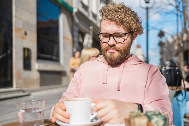 Milenium brodaty człowiek jedzący śniadanie przy stole ulicznej kawiarni w wiosenny dzień, pijący ciepłe cappuccino. Wiosenny taras restauracji. - Zdjęcie, obraz