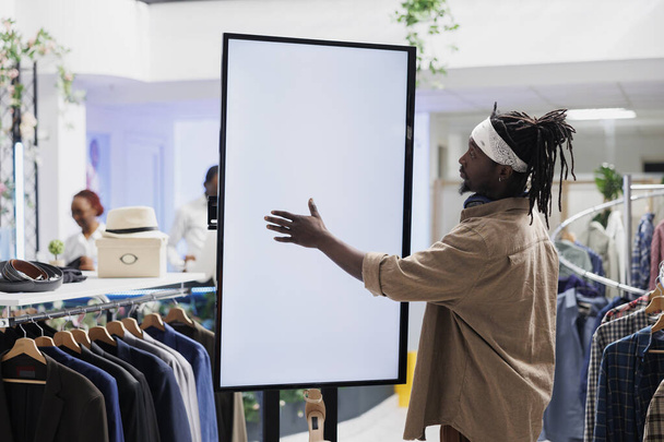 A vásárló üres fehér okos képernyőt használ a ruhák böngészéséhez. Afrikai amerikai stílusos férfi megérinti interaktív üres kijelző vásárlás közben ruházat plázában butik - Fotó, kép
