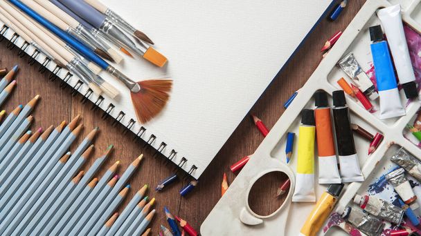 Draufsicht auf Holztisch mit Bleistiften, Farbtuben, Kunstzubehör, verschiedenen Pinseln auf weißem Papier - Foto, Bild