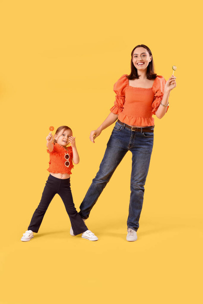 Όμορφη μητέρα και χαριτωμένη μικρή κόρη της με γλυκά γλειφιτζούρια σε κίτρινο φόντο - Φωτογραφία, εικόνα