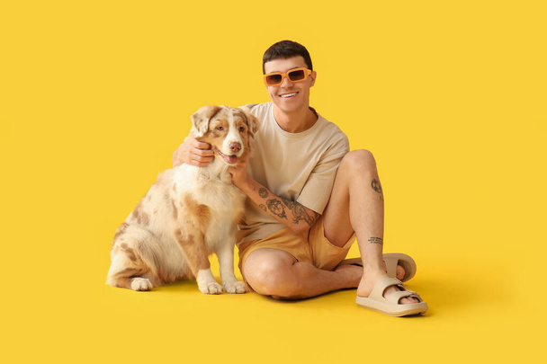 Giovane uomo in occhiali da sole con cane pastore australiano seduto su sfondo giallo - Foto, immagini
