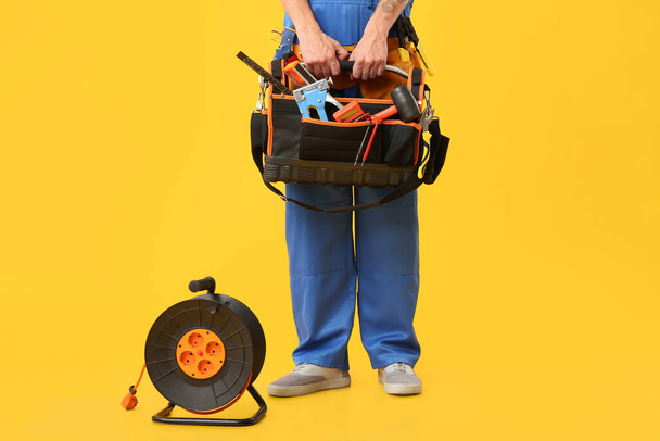 Electricista masculino con bolsa de herramientas y carrete de cable de extensión sobre fondo amarillo - Foto, imagen