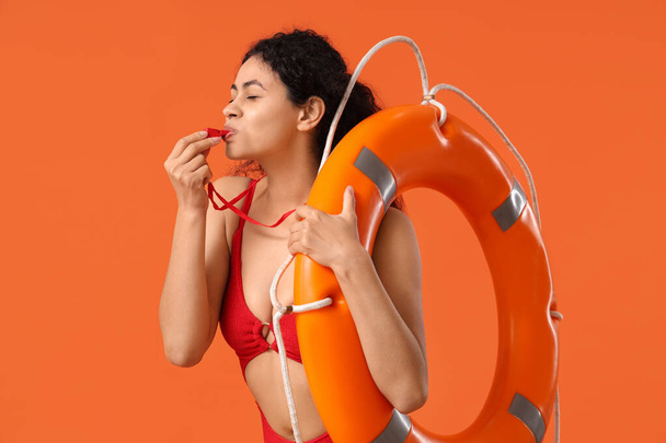 Schöne junge afroamerikanische Rettungsschwimmerin mit Ringboje, die auf orangefarbenem Hintergrund pfeift - Foto, Bild
