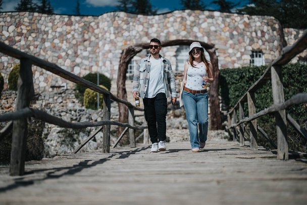 Um jovem casal alegre caminha de mãos dadas através de uma ponte de madeira encantadora, incorporando o espírito de férias e explorando novos lugares juntos. - Foto, Imagem