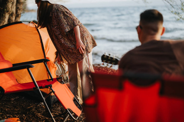 キャンプ旅行中に湖の近くでリラックスする2人の友人,オレンジ色のテントとギター,自然と他社を楽しむ. - 写真・画像